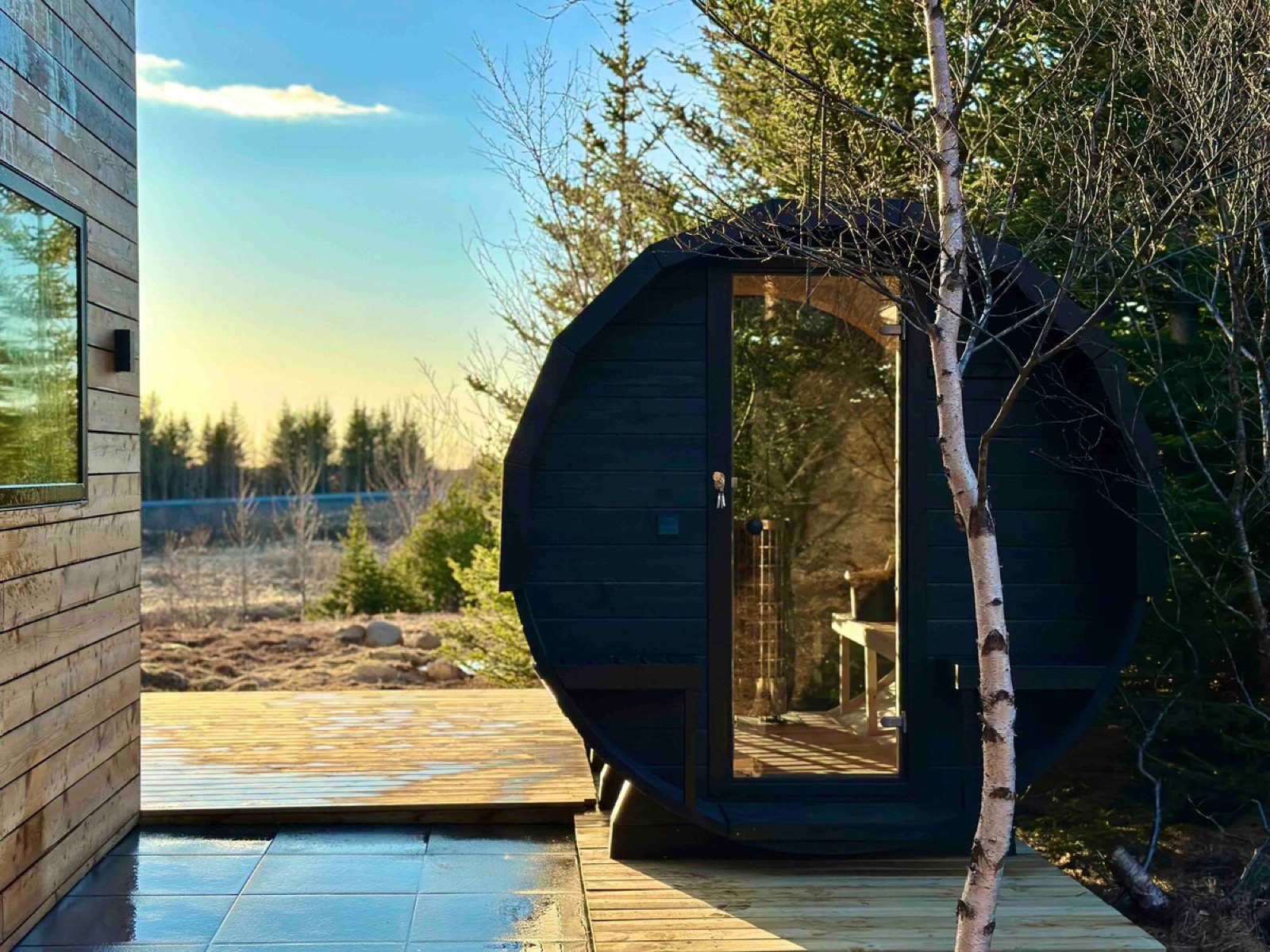 The black barrel sauna on the deck at sunset, its round window lit