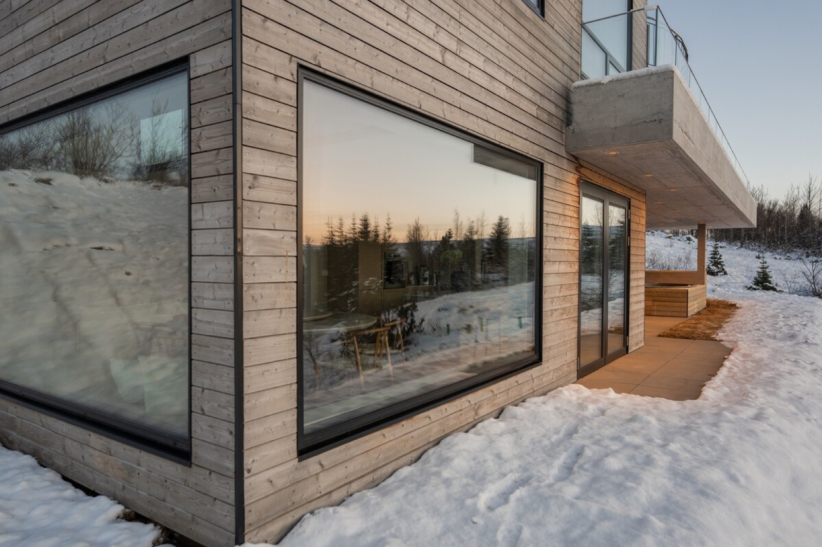 The timber cladding meeting its concrete plinth, a wide window reflecting the trees